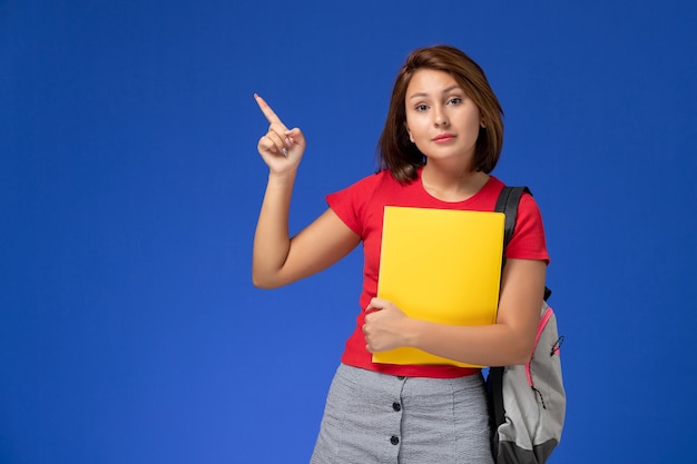 Vue de face jeune étudiante en chemise rouge avec sac à dos tenant des fichiers jaunes sur le fond bleu clair.