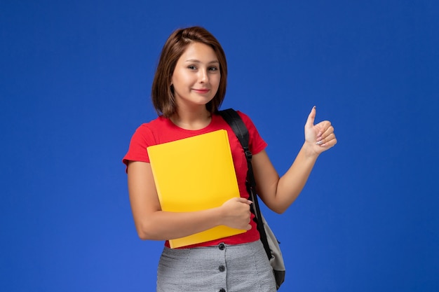 Vue de face jeune étudiante en chemise rouge avec sac à dos tenant des fichiers jaunes sur le fond bleu clair.