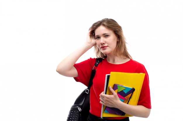 Une vue de face jeune étudiante en chemise rouge sac noir tenant un stylo et des cahiers sur le blanc