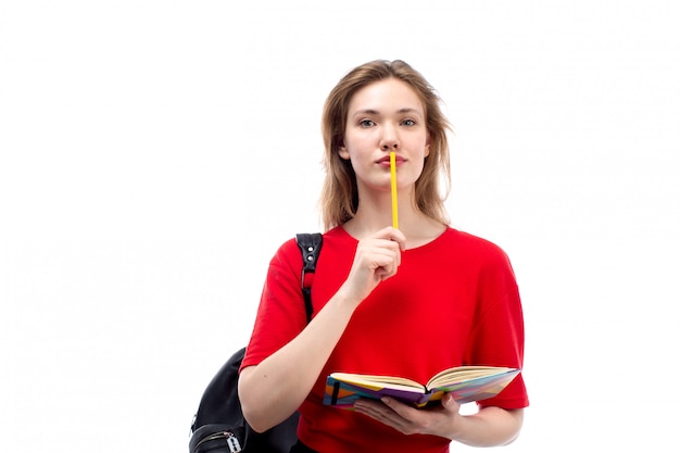 Une vue de face jeune étudiante en chemise rouge sac noir tenant un stylo et des cahiers sur le blanc