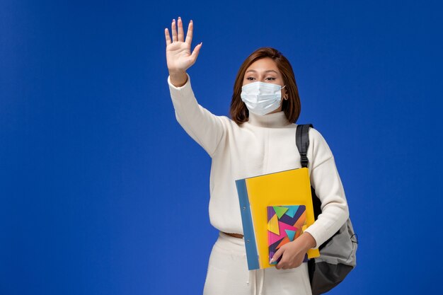 Vue de face jeune étudiante en maillot blanc portant un masque avec sac et cahier en agitant sur le mur bleu