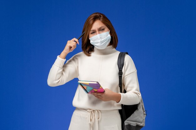 Vue de face jeune étudiante en maillot blanc portant un masque avec sac et cahier sur mur bleu