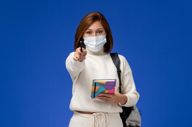 Vue de face jeune étudiante en maillot blanc portant un masque avec sac et cahier avec stylo sur le mur bleu