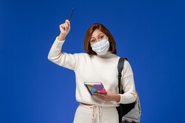 Vue de face jeune étudiante en maillot blanc portant un masque avec sac et cahier avec stylo sur le mur bleu