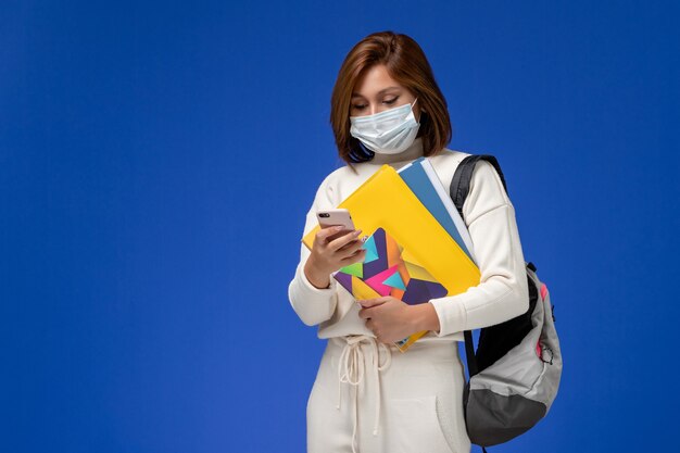 Vue de face jeune étudiante en maillot blanc portant un masque avec sac et cahiers et à l'aide de son téléphone sur le mur bleu