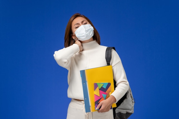 Vue de face jeune étudiante en maillot blanc portant un masque avec sac et cahiers ayant mal au cou sur le mur bleu