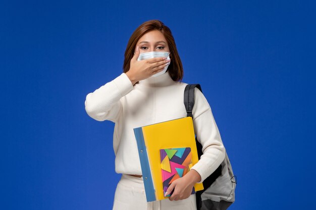 Vue de face jeune étudiante en maillot blanc portant un masque avec sac et cahiers fermant sa bouche sur le mur bleu