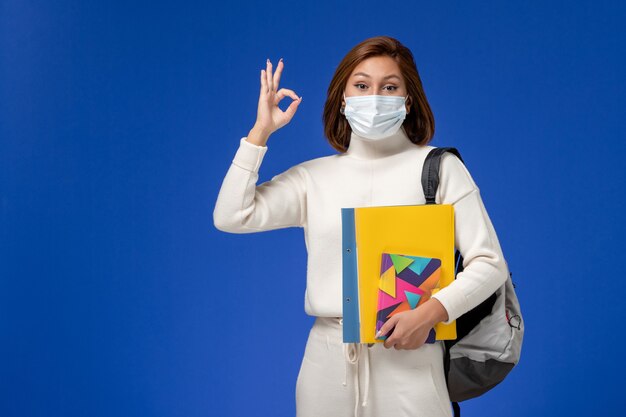 Vue de face jeune étudiante en maillot blanc portant un masque avec sac et cahiers sur le mur bleu