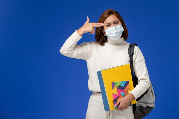 Vue de face jeune étudiante en maillot blanc portant un masque avec sac et cahiers sur le mur bleu