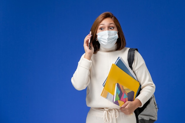 Vue de face jeune étudiante en maillot blanc portant un masque avec sac et cahiers de parler au téléphone sur mur bleu