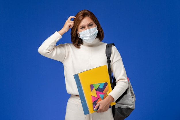 Vue de face jeune étudiante en maillot blanc portant un masque avec sac et cahiers pensant sur le mur bleu