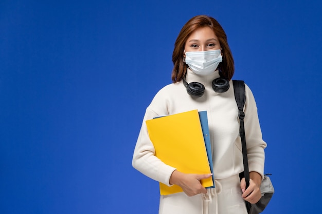 Vue de face jeune étudiante en maillot blanc portant un masque et un sac à dos tenant des fichiers sur le mur bleu