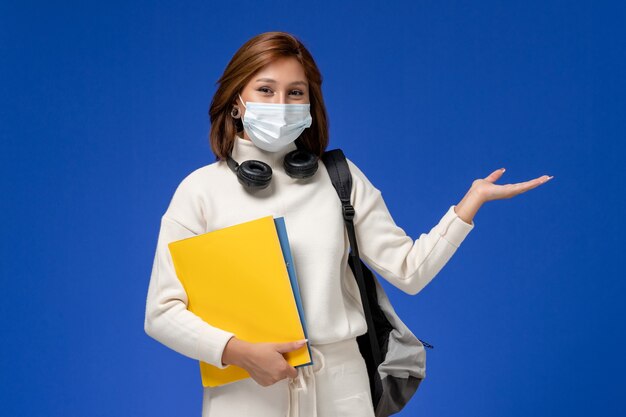 Vue de face jeune étudiante en maillot blanc portant un masque et un sac à dos tenant des fichiers sur le mur bleu