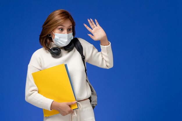 Vue de face jeune étudiante en maillot blanc portant un masque et un sac à dos tenant des fichiers sur le mur bleu