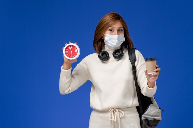 Vue de face jeune étudiante en maillot blanc portant un masque et un sac à dos tenant horloge et café sur mur bleu