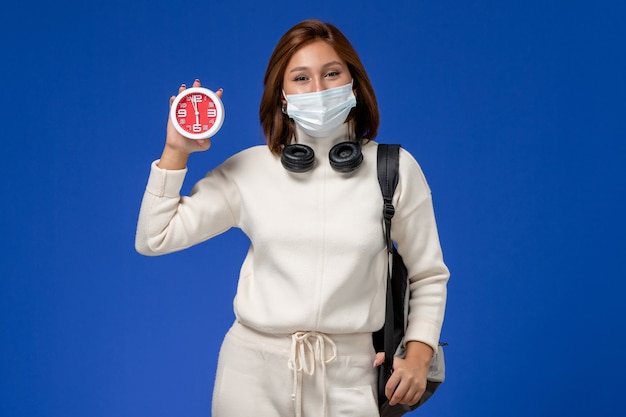 Vue de face jeune étudiante en maillot blanc portant un masque et un sac à dos tenant horloge sur mur bleu