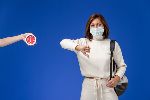 Vue de face jeune étudiante en maillot blanc portant un masque et un sac montrant un signe différent sur le mur bleu