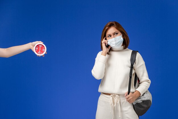 Vue de face jeune étudiante en maillot blanc portant un masque et un sac pensant sur le mur bleu