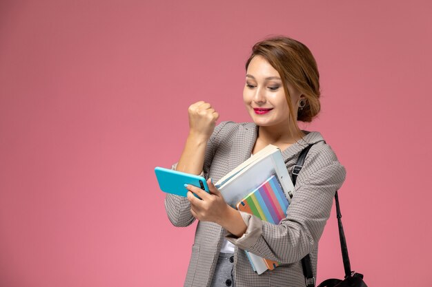 Vue de face jeune étudiante en manteau gris avec des cahiers et à l'aide d'un téléphone avec le sourire sur les leçons de fond rose étude de collège universitaire