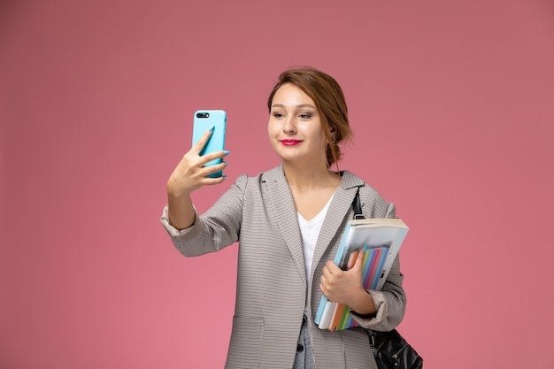 Vue de face jeune étudiante en manteau gris avec des cahiers en prenant un selfie sur fond rose leçons college college study