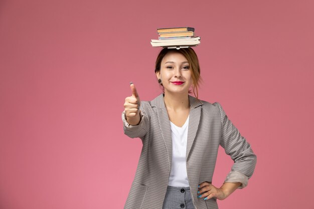 Vue de face jeune étudiante en manteau gris avec des cahiers sur sa tête souriant sur fond rose leçons étude de collège universitaire