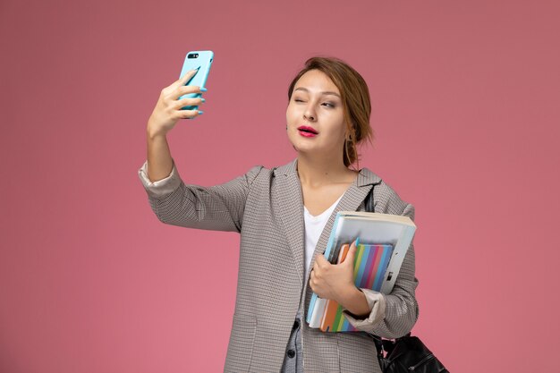 Vue de face jeune étudiante en manteau gris posant et prenant un selfie sur fond rose leçons étude de collège universitaire