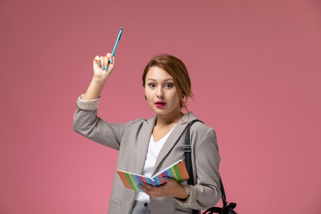 Vue de face jeune étudiante en manteau gris posant tenant un cahier avec un stylo surélevé sur le fond rose leçons université college