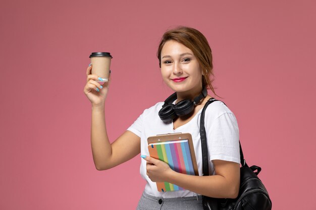 Vue de face jeune étudiante en t-shirt blanc avec cahier et sac posant et tenant une tasse de café sur fond rose leçon université cahier d'étude