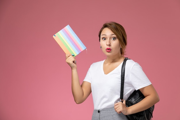 Vue de face jeune étudiante en t-shirt blanc et pantalon gris avec cahier dans ses mains sur fond rose leçons étudiantes college college