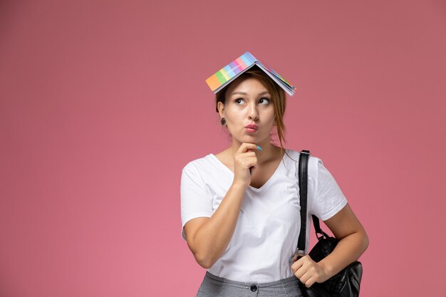 Vue de face jeune étudiante en t-shirt blanc et pantalon gris avec cahier sur sa tête et expression de la pensée sur le bureau rose leçons college college