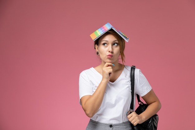 Vue de face jeune étudiante en t-shirt blanc et pantalon gris avec cahier sur sa tête et expression de la pensée sur le bureau rose leçons college college