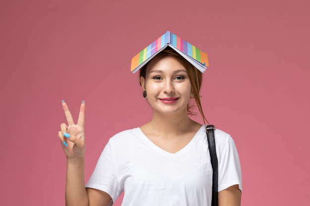 Vue de face jeune étudiante en t-shirt blanc et pantalon gris avec cahier sourire et geste de victoire sur fond rose leçons étudiantes college college