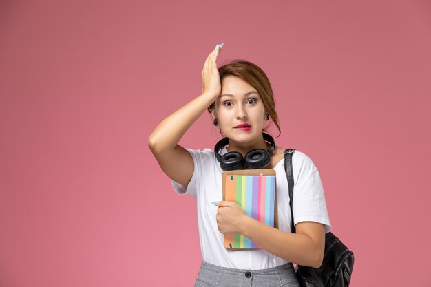 Vue de face jeune étudiante en t-shirt blanc et pantalon gris avec des écouteurs et un cahier dans ses mains sur fond rose leçons étudiantes college college