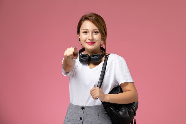 Vue de face jeune étudiante en t-shirt blanc et pantalon gris avec écouteurs souriant sur fond rose leçons étudiants college college