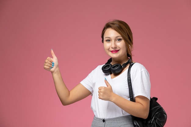 Vue de face jeune étudiante en t-shirt blanc et pantalon gris avec écouteurs et sourire sur fond rose cours étudiants college college