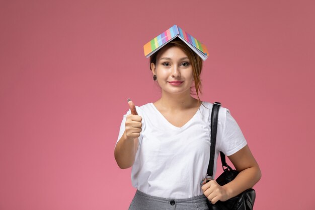 Vue de face jeune étudiante en t-shirt blanc et pantalon gris souriant sur le fond rose leçons étudiantes college college