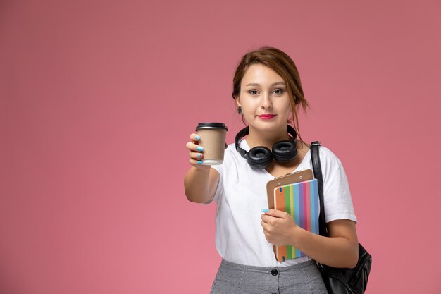 Vue de face jeune étudiante en t-shirt blanc avec sac et écouteurs posant et souriant tenant un café sur le livre d'étude de l'université de la leçon de fond rose