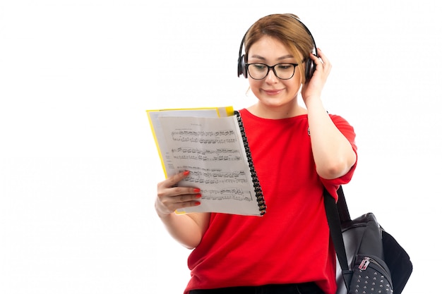 Une vue de face jeune étudiante en t-shirt rouge jeans noirs écouter de la musique à travers des écouteurs noirs lire des notes sur le blanc