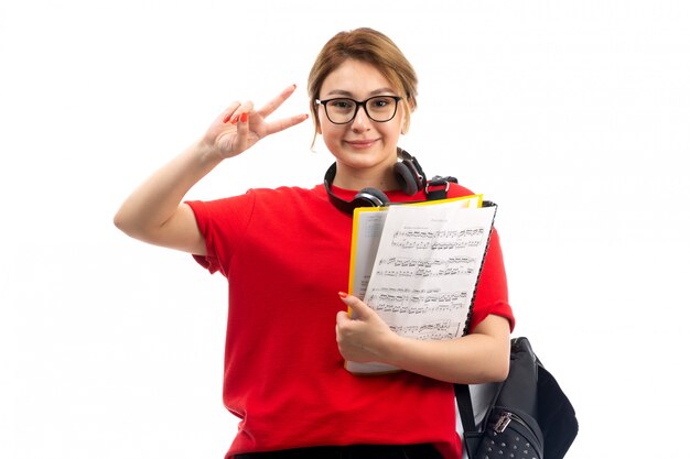 Une vue de face jeune étudiante en t-shirt rouge jeans noirs portant des écouteurs noirs tenant un cahier de notes souriant sur le blanc