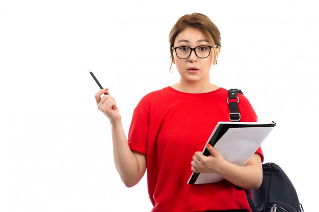 Une vue de face jeune étudiante en t-shirt rouge jeans noirs tenant un cahier sur le blanc