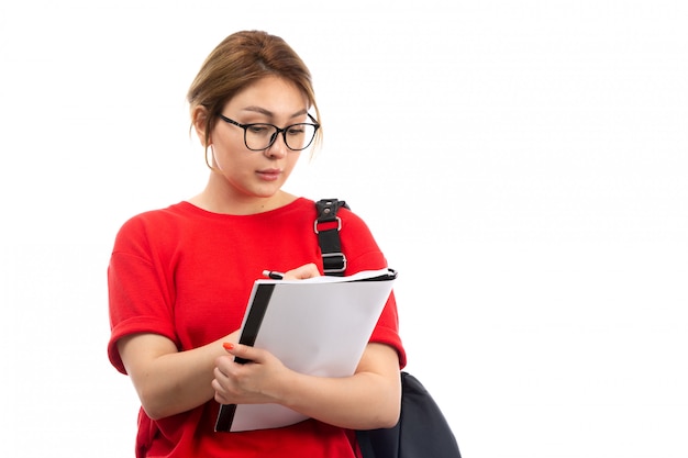 Une vue de face jeune étudiante en t-shirt rouge jeans noirs tenant différents cahiers et fichiers en prenant des notes sur le blanc