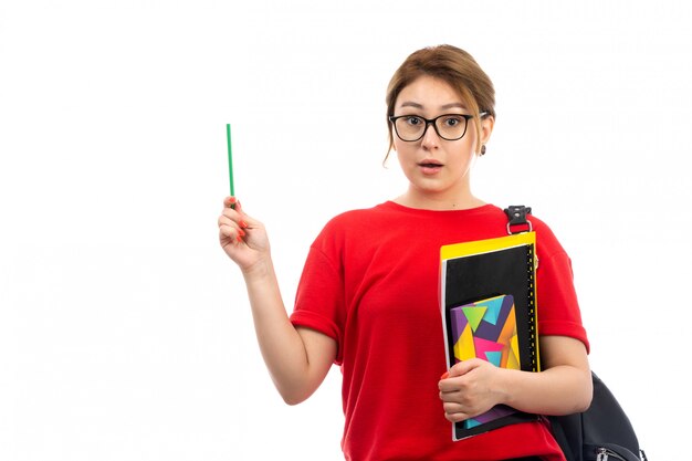 Une vue de face jeune étudiante en t-shirt rouge jeans noirs tenant différents cahiers et fichiers tenant un crayon avec sac sur le blanc