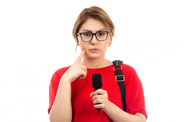 Une vue de face jeune étudiante en t-shirt rouge portant un sac noir tenant l'expression d'hésitant microphone sur le blanc