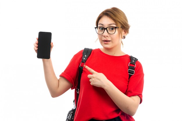 Une Vue De Face Jeune étudiante En T-shirt Rouge Portant Un Sac Noir Tenant Un Smartphone Noir Sur Le Blanc