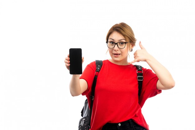 Une vue de face jeune étudiante en t-shirt rouge portant un sac noir tenant un smartphone noir souriant sur le blanc