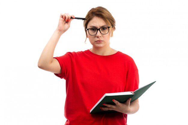 Une vue de face jeune étudiante en t-shirt rouge tenant un cahier d'écrire des notes de réflexion sur le blanc