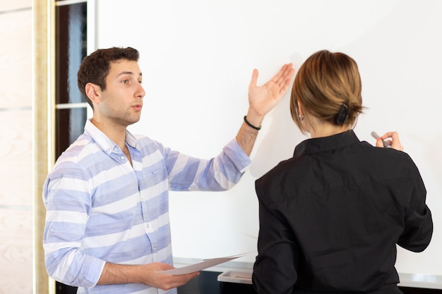 Photo gratuite une vue de face jeune femme d'affaires attrayant en chemise noire avec un jeune homme discutant des graphiques sur le bureau pendant que la jeune femme présente son travail en lisant le document de présentation de la création d'emploi