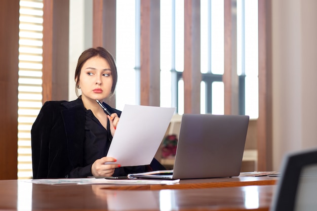 Une vue de face jeune femme d'affaires attrayant en chemise noire veste noire à l'aide de son ordinateur portable argent écrit lecture travaillant à l'intérieur de son travail de bureau bâtiment de l'emploi