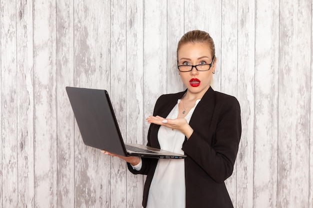 Vue de face jeune femme d'affaires dans des vêtements stricts veste noire à l'aide d'un ordinateur portable sur un bureau blanc