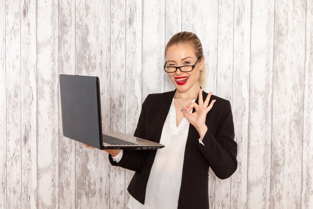 Vue de face jeune femme d'affaires dans des vêtements stricts veste noire à l'aide de son ordinateur portable un clin de œil sur le mur blanc travail travail bureau femme travailleur